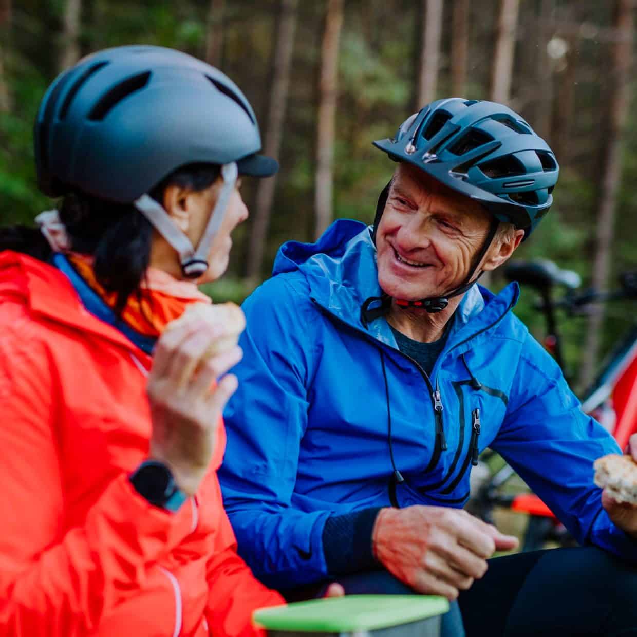 Older couple wearing cycling helmets and outdoor jackets, sitting in a forest having a snack break with their bicycles nearby.
