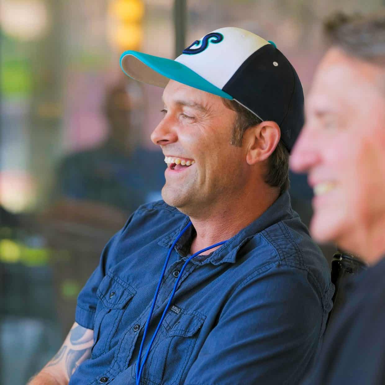 Two men sitting and smiling in conversation; one is wearing a baseball cap and a blue shirt, with a blurred indoor background.