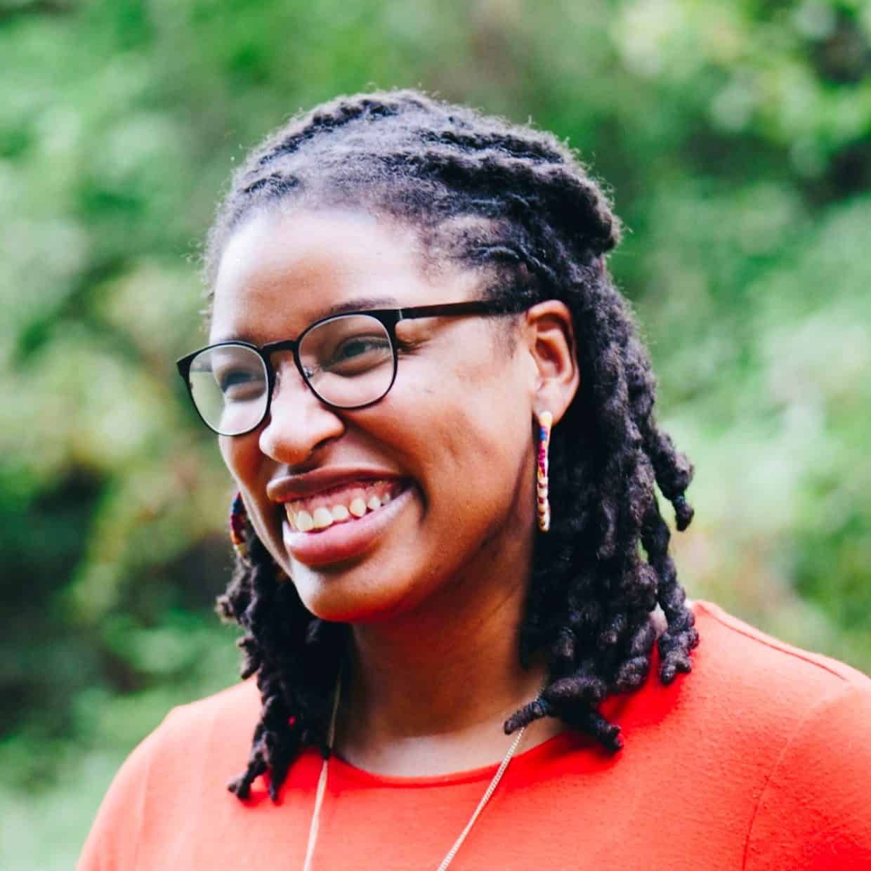 Smiling woman in red top with glasses and earrings standing outdoors.