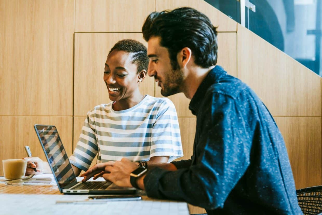 Two colleagues smiling while working together at a laptop in a modern office setting, with a coffee cup and notepad on the table.