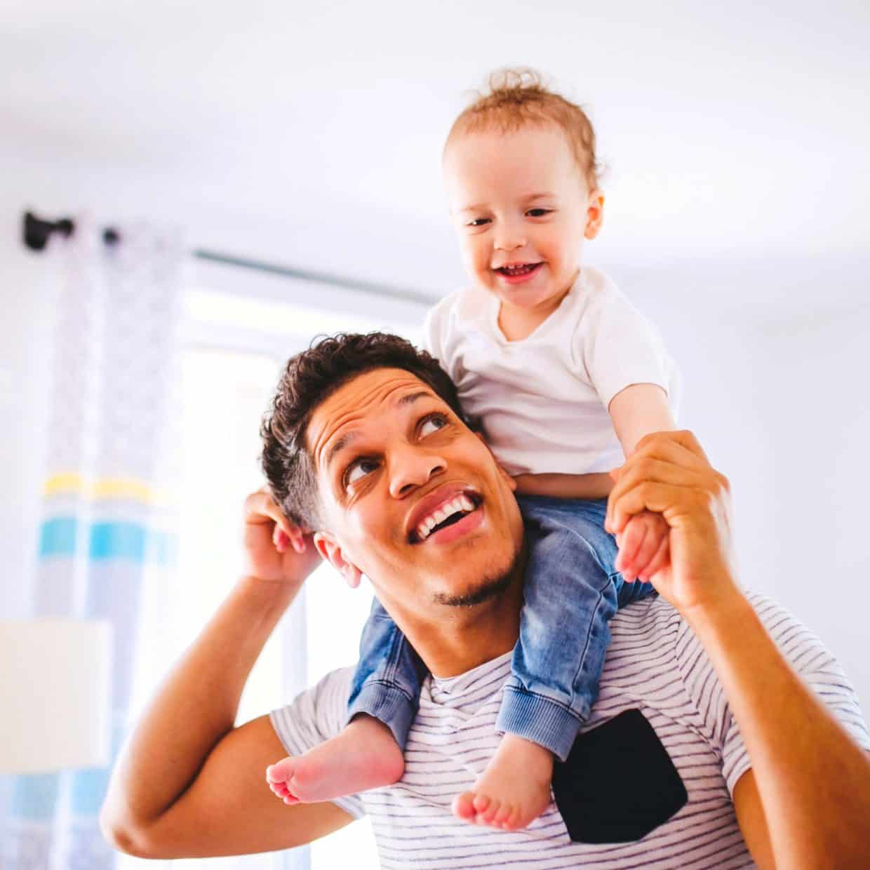 Smiling father holding his young child on his shoulders in a bright living room.