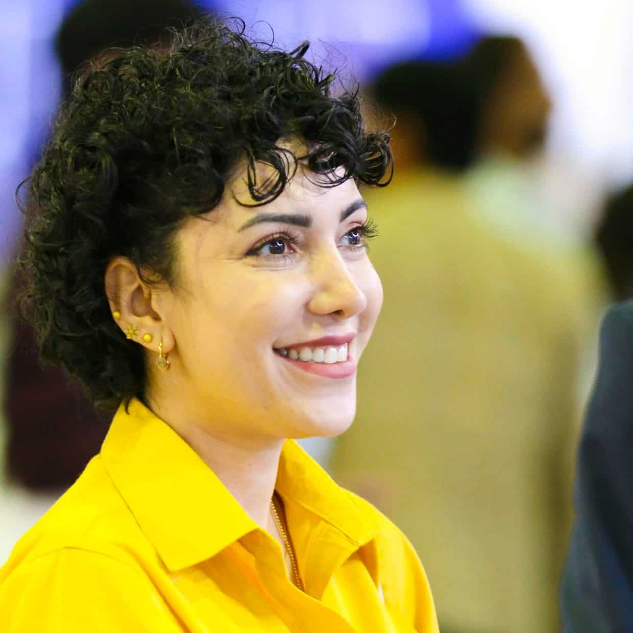 Smiling woman in a yellow shirt at an indoor event with people in the background.