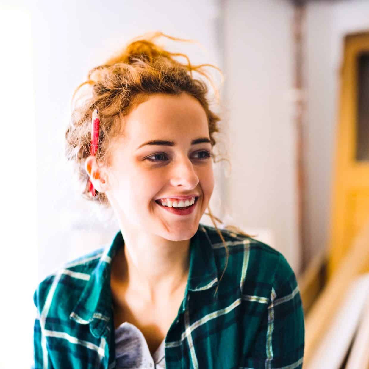 Smiling woman in a plaid shirt with a pencil in her hair standing in a workshop.