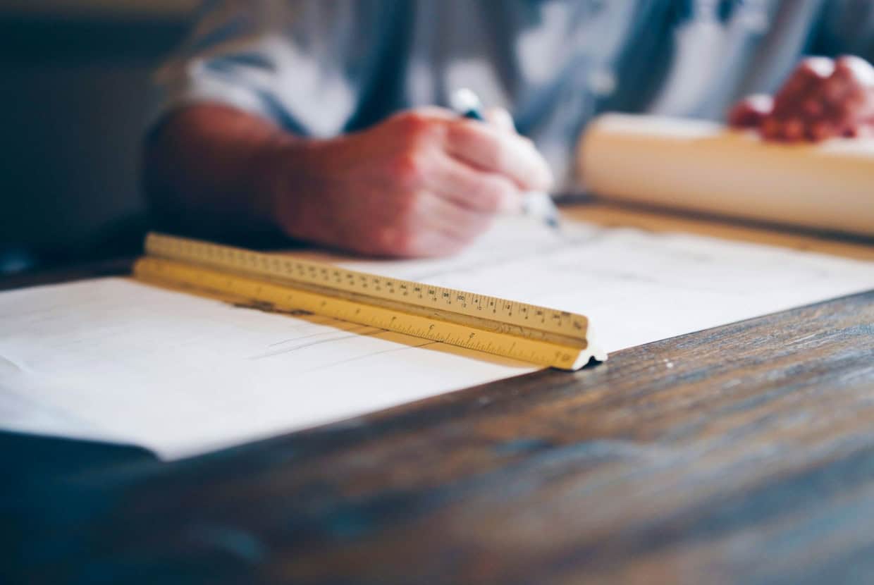 Architect working on building plans at a desk with a yellow ruler and pen in hand.