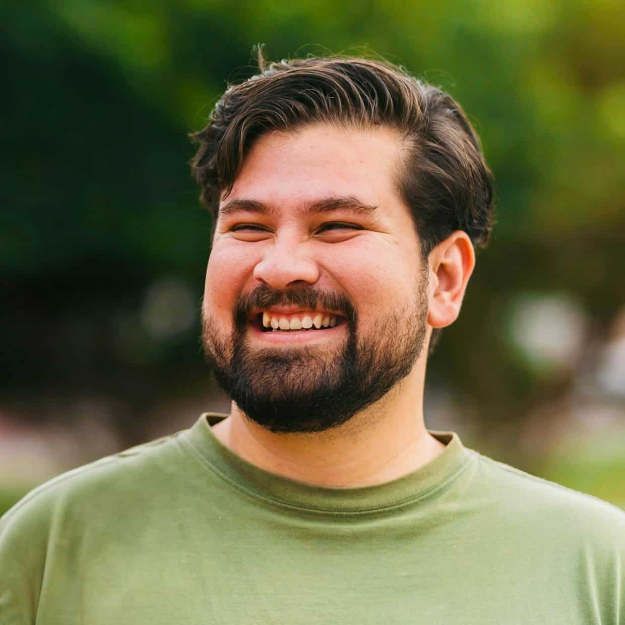 Man with beard in a green shirt smiling outdoors with trees in the background.