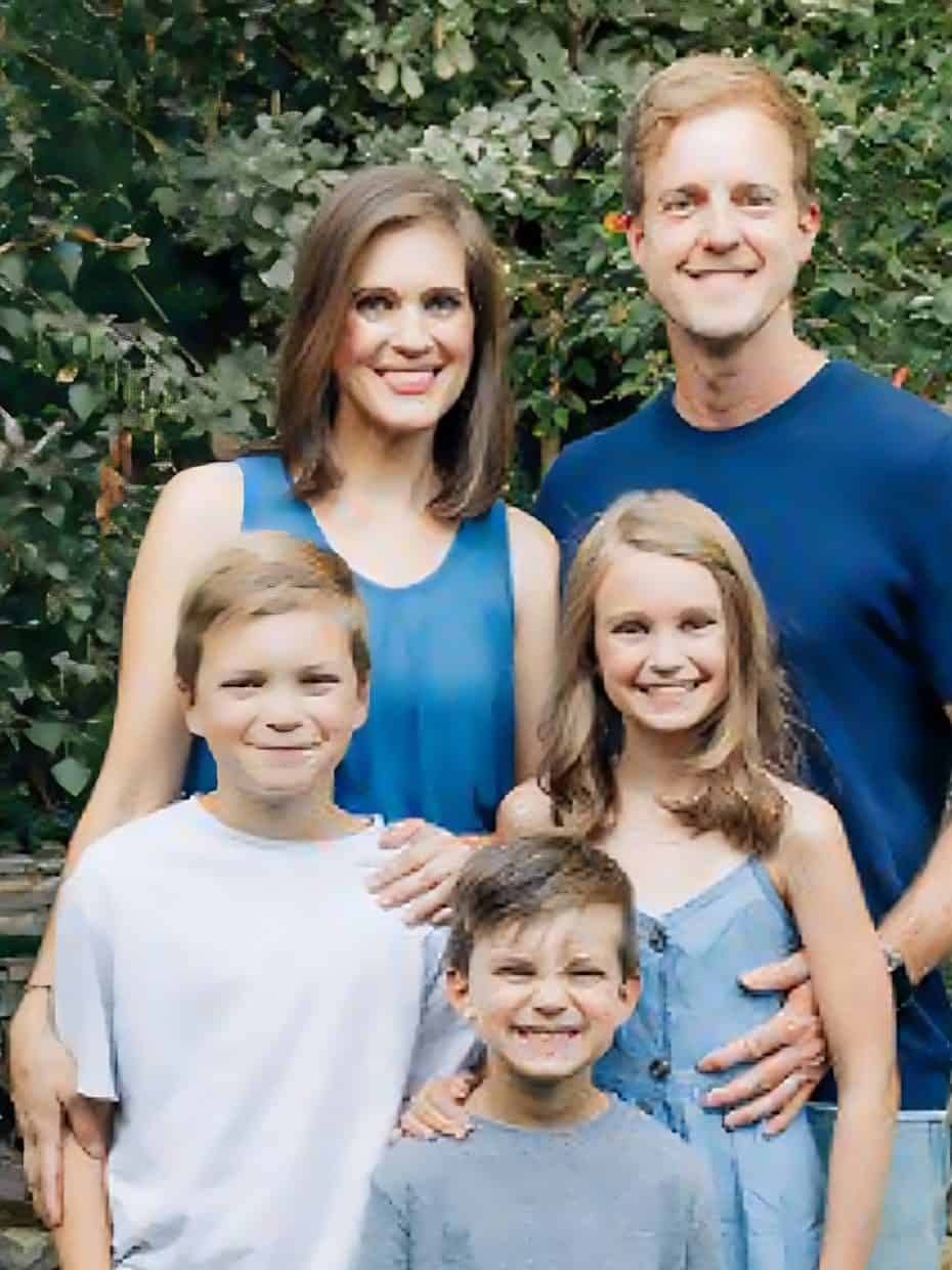 Smiling family of five posing outdoors in front of green foliage, with parents standing behind and three children in front.