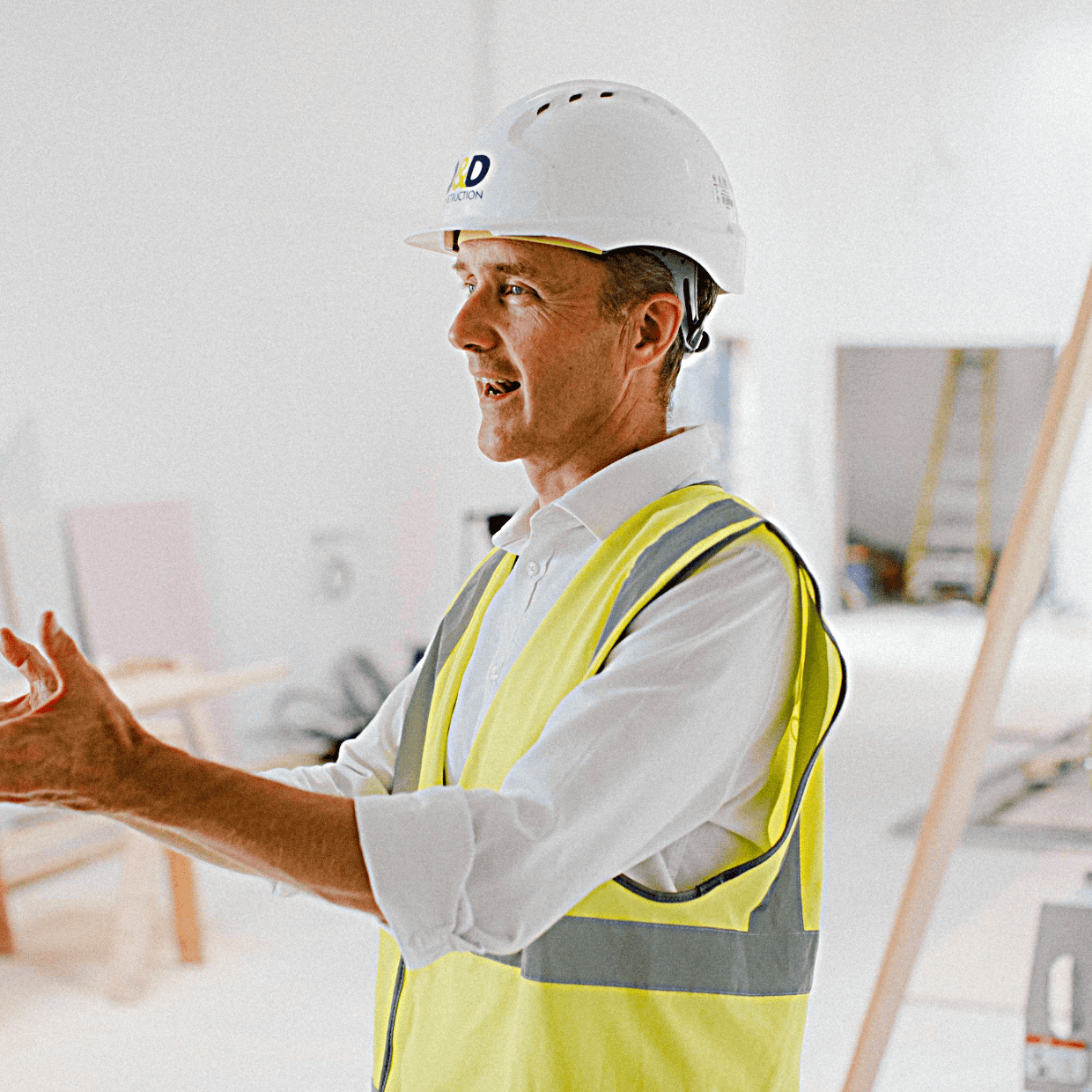 Man wearing a hard hat and high visibility vest gesturing while speaking inside a building under construction with tools and equipment in the background.