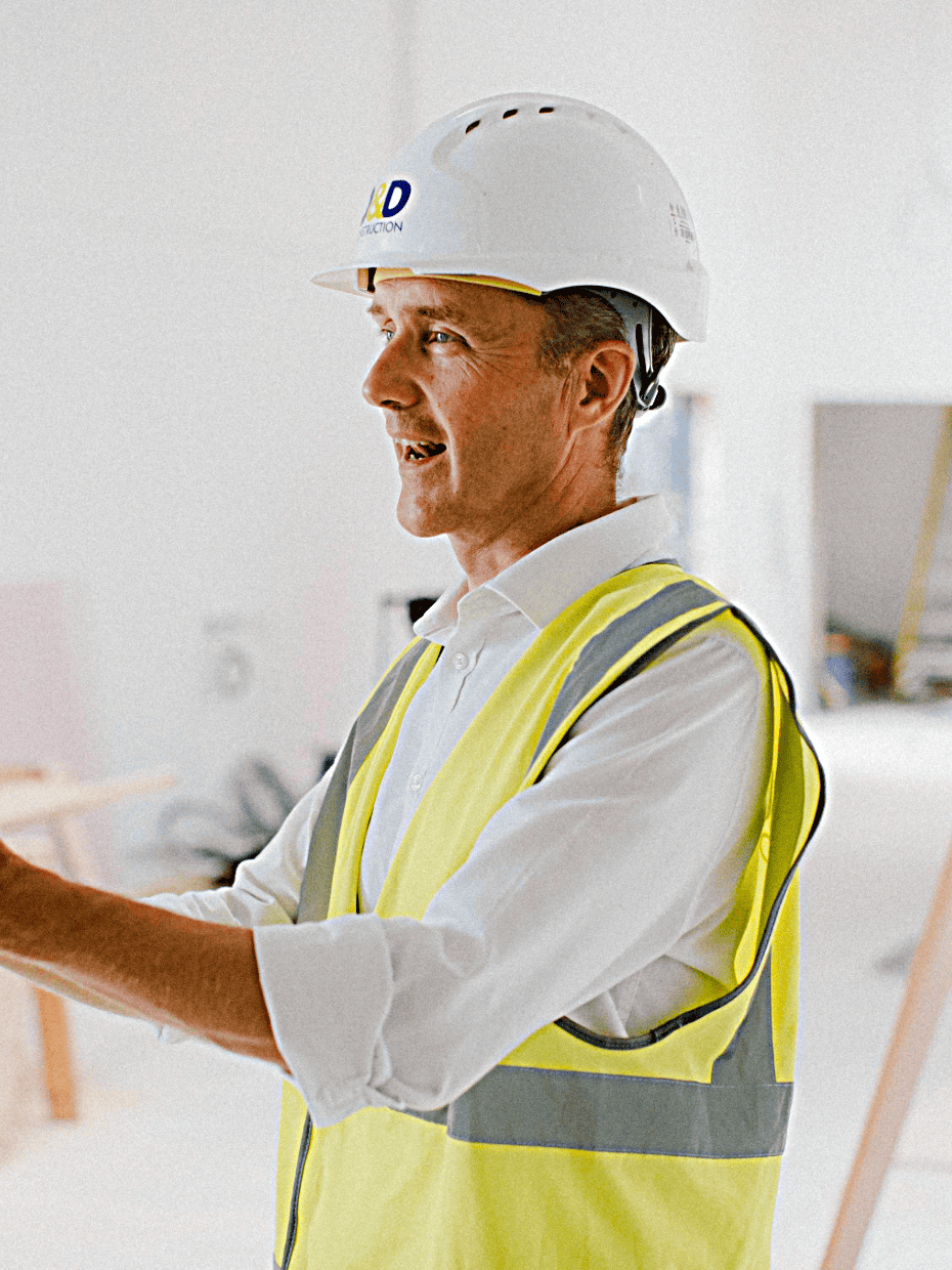 Man wearing a hard hat and high visibility vest gesturing while speaking inside a building under construction with tools and equipment in the background.