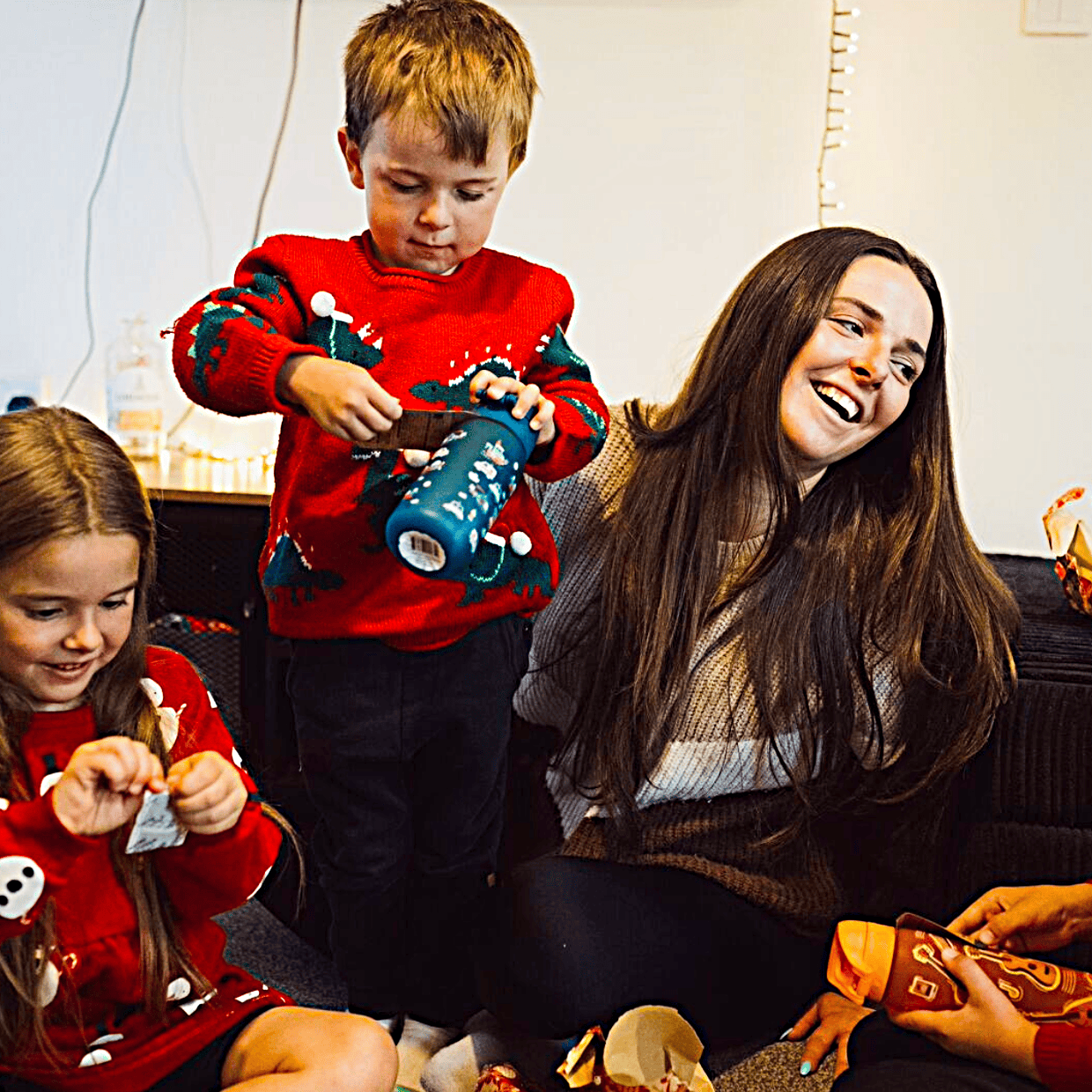 Mother and three children wearing festive jumpers sitting by a Christmas tree, smiling and opening presents together at home.