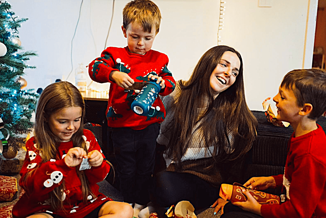 Mother and three children wearing festive jumpers sitting by a Christmas tree, smiling and opening presents together at home.