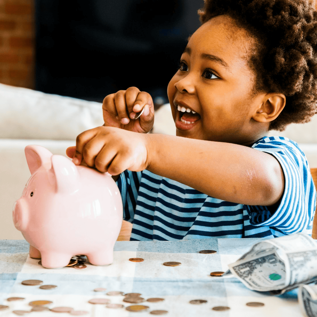 Young child smiling while placing coins into a pink piggy bank, with loose change on the table, representing early saving habits and financial learning.