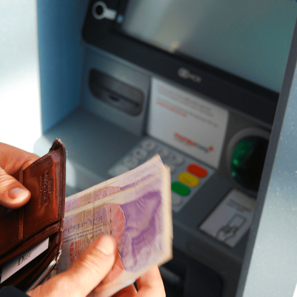 Person holding a wallet and banknotes while standing at an ATM machine, with card slots and keypad visible in the background.