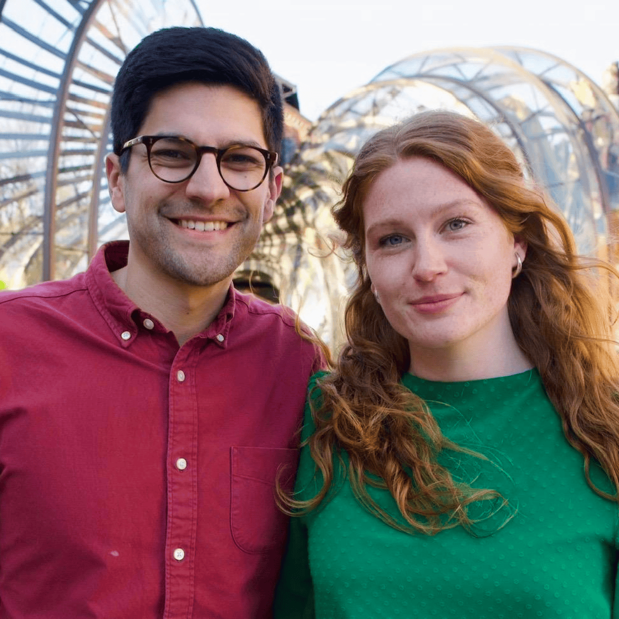 John and Imogen standing side by side outdoors, smiling at the camera with a modern arched structure and buildings in the background.