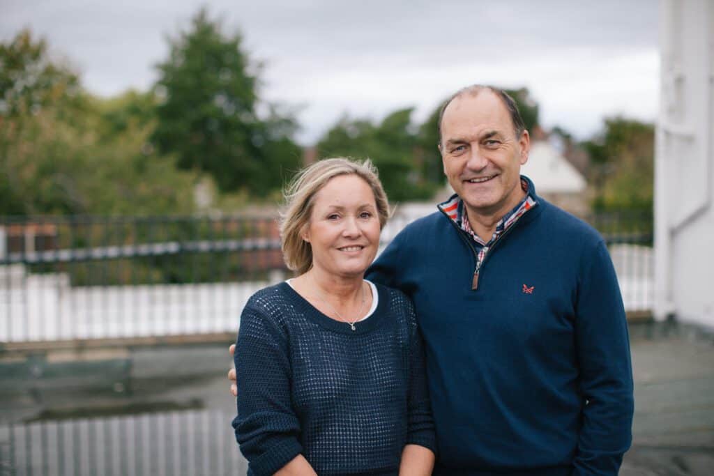 Smiling couple standing outdoors together on a cloudy day, with trees and houses in the background.