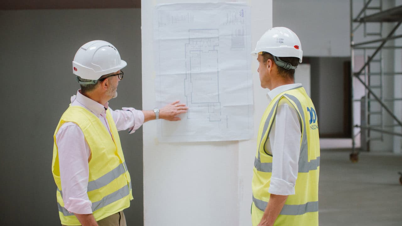 Two construction professionals wearing hard hats and high visibility vests reviewing architectural plans on a wall inside a building site.