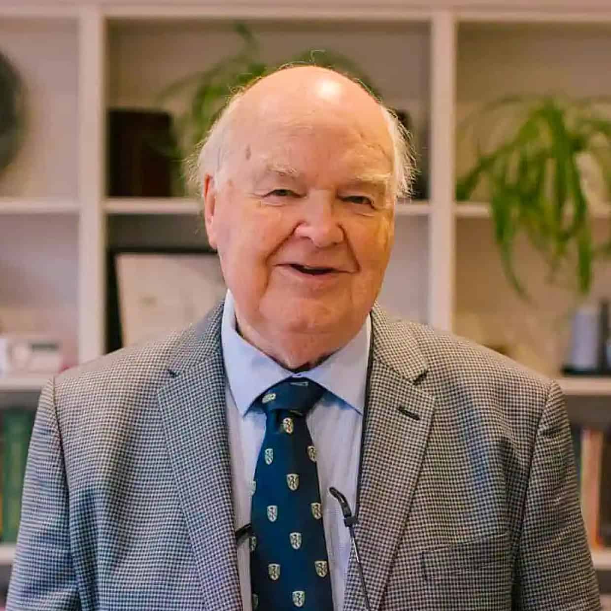 John Lennox in a grey checkered suit and blue tie smiling while standing in front of a bookshelf filled with books and plants.