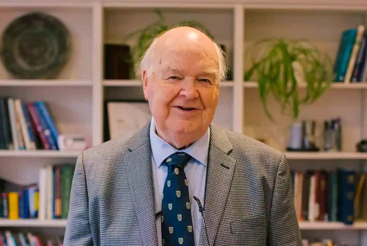 John Lennox in a grey checkered suit and blue tie smiling while standing in front of a bookshelf filled with books and plants.