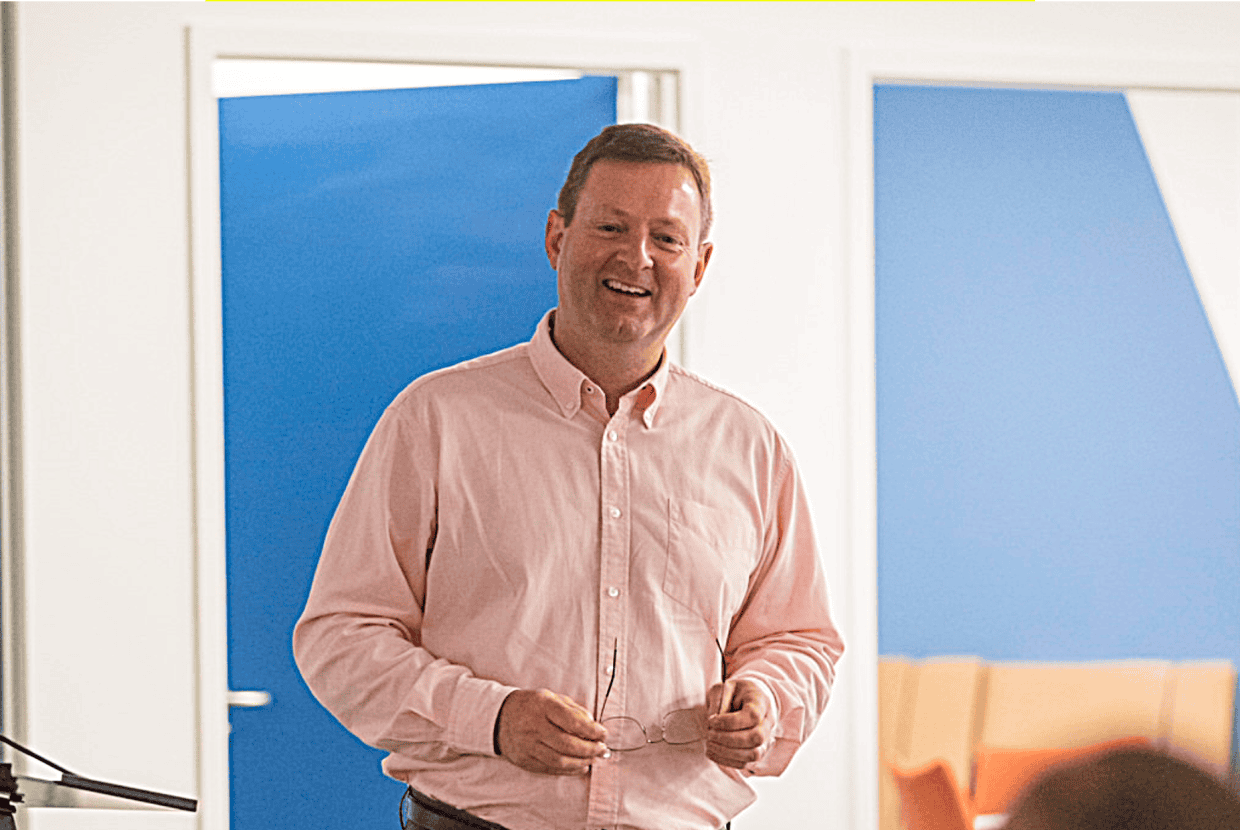 Paul Houghton smiling while speaking indoors, holding glasses in his hands with a bright background behind him.