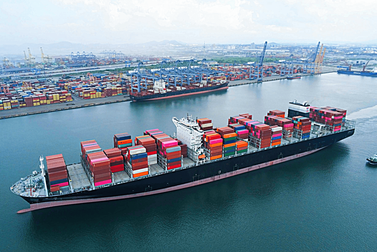 Large container ship carrying multicoloured cargo containers sailing past a busy commercial port with cranes and stacked containers.