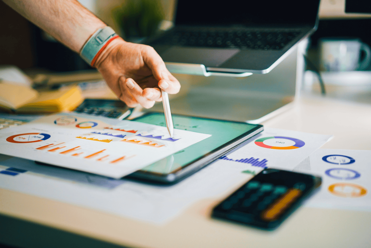 Hand using a stylus on a tablet displaying charts and graphs, with printed financial reports, calculator, and laptop on a desk.