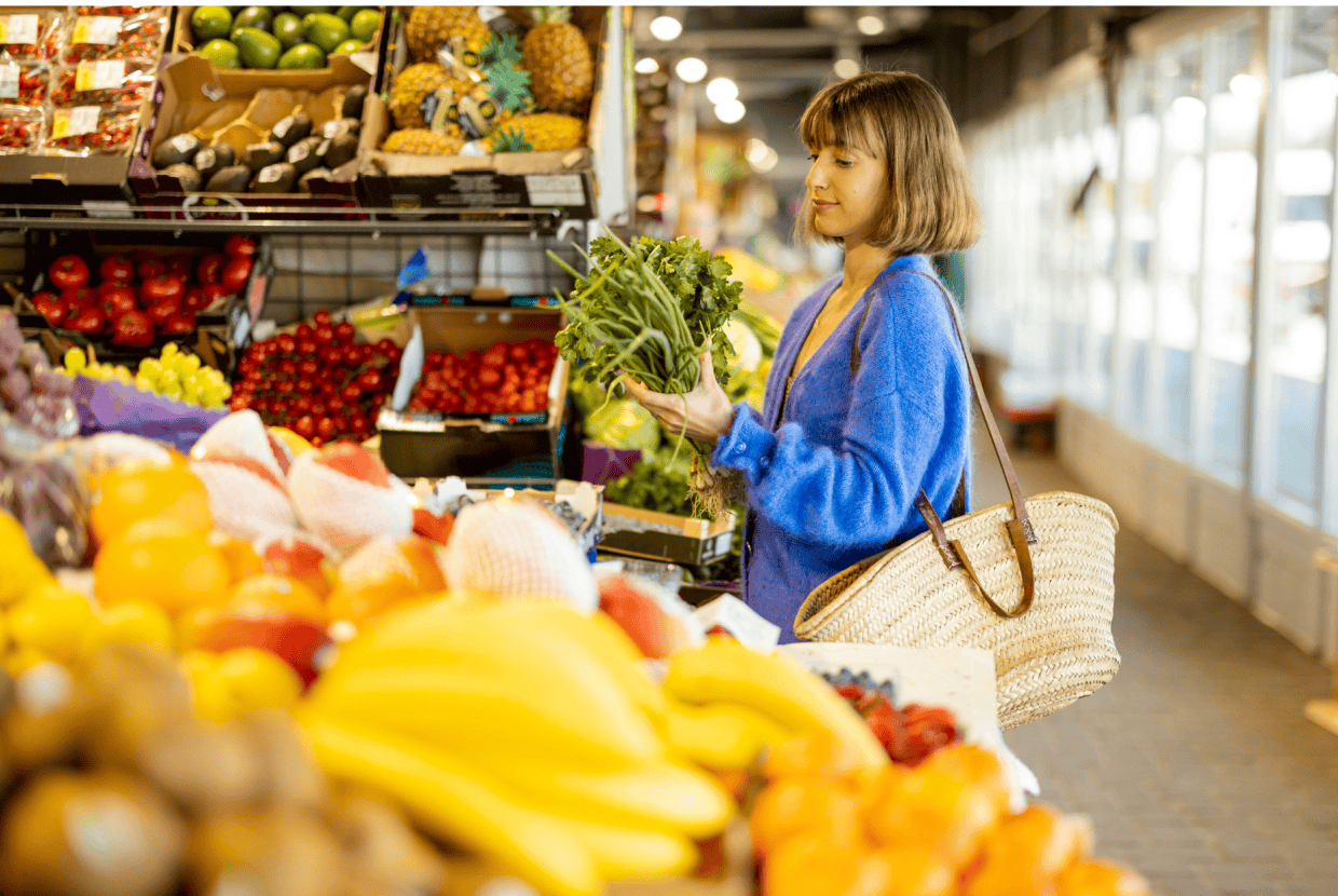 Woman selecting fresh vegetables at a market stall with fruit and produce displayed around her.