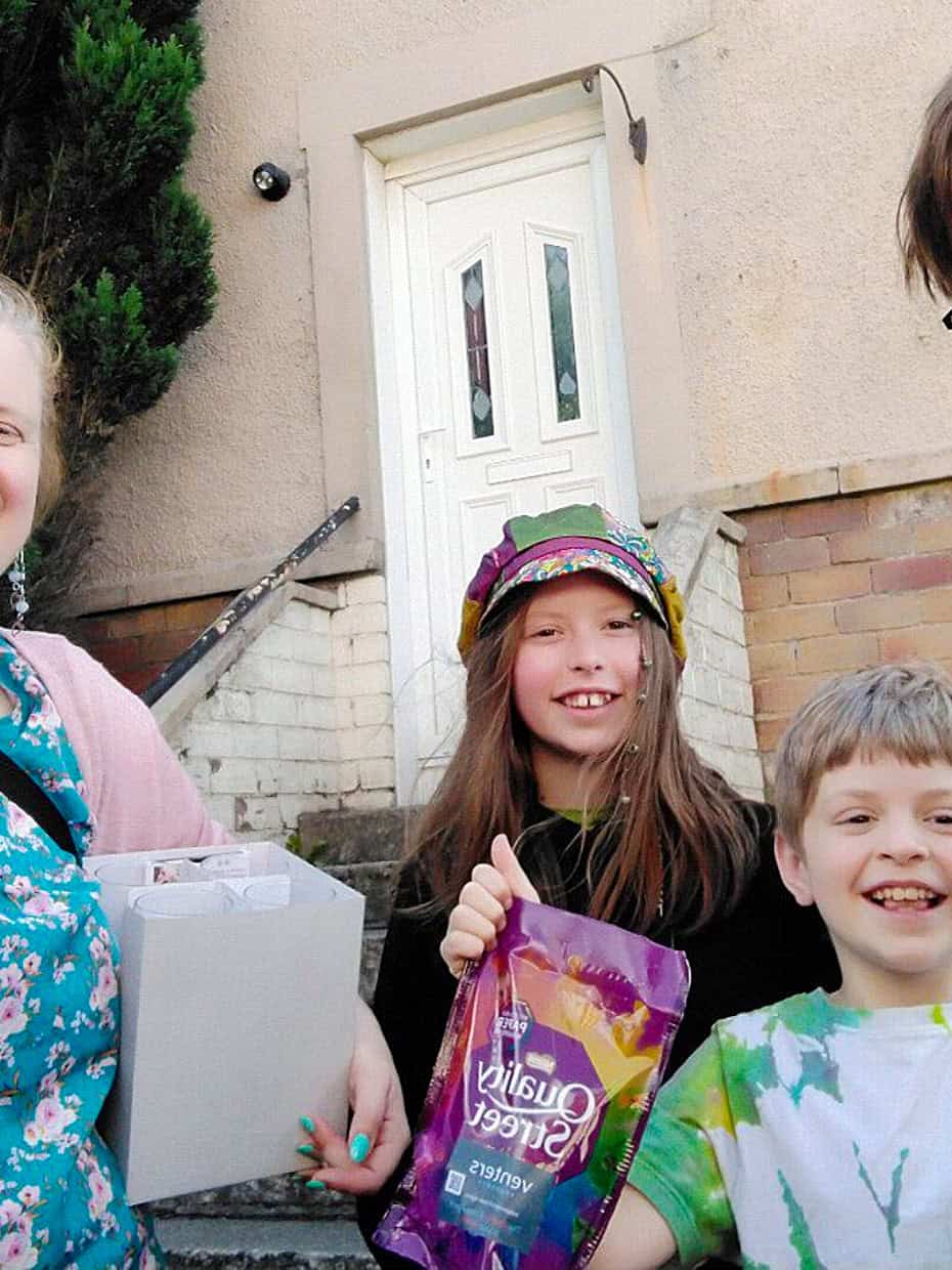 Smiling family of four standing outside their home, with the mother holding a box and the children holding sweets, celebrating with excitement.