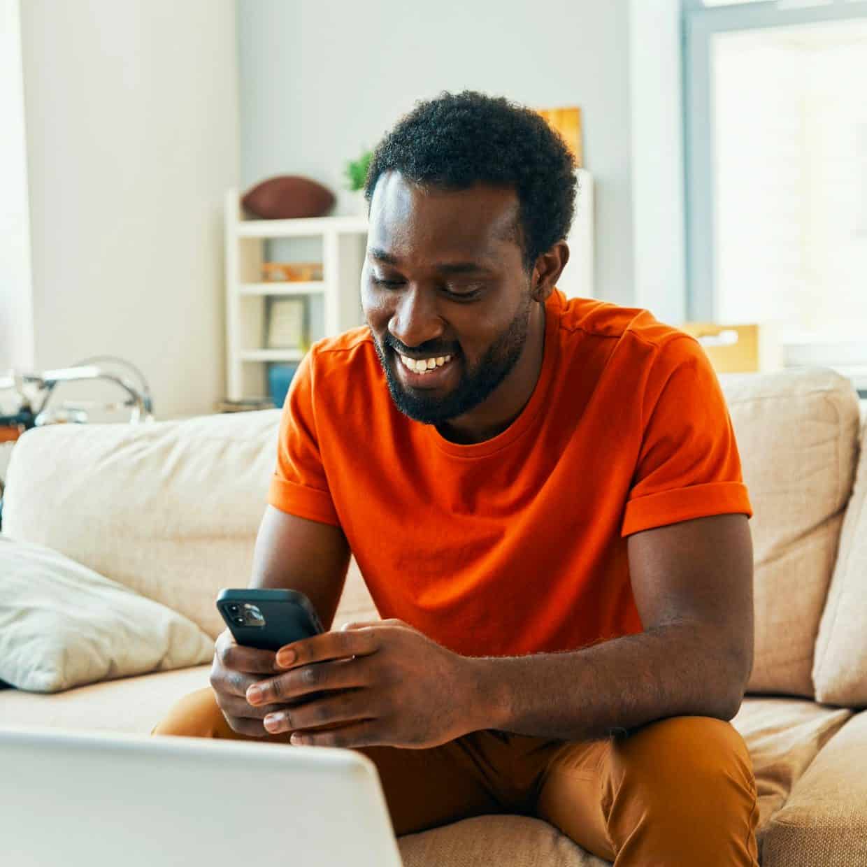 Smiling man sitting on a sofa using his phone with a laptop in front of him.