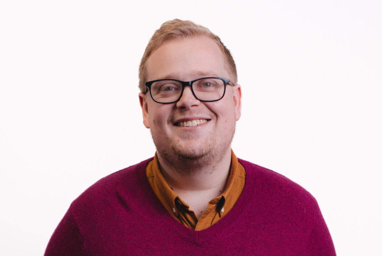 Smiling man wearing glasses, a mustard shirt, and a burgundy jumper, standing against a plain white background.
