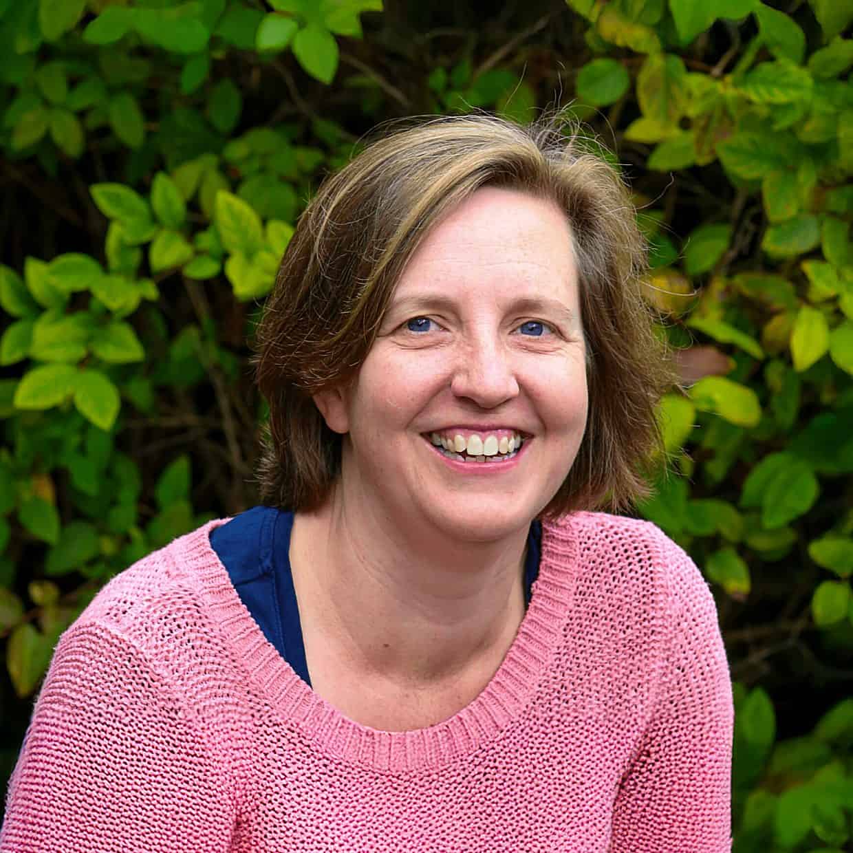 Smiling woman wearing a pink jumper sitting outdoors in front of green leafy bushes.
