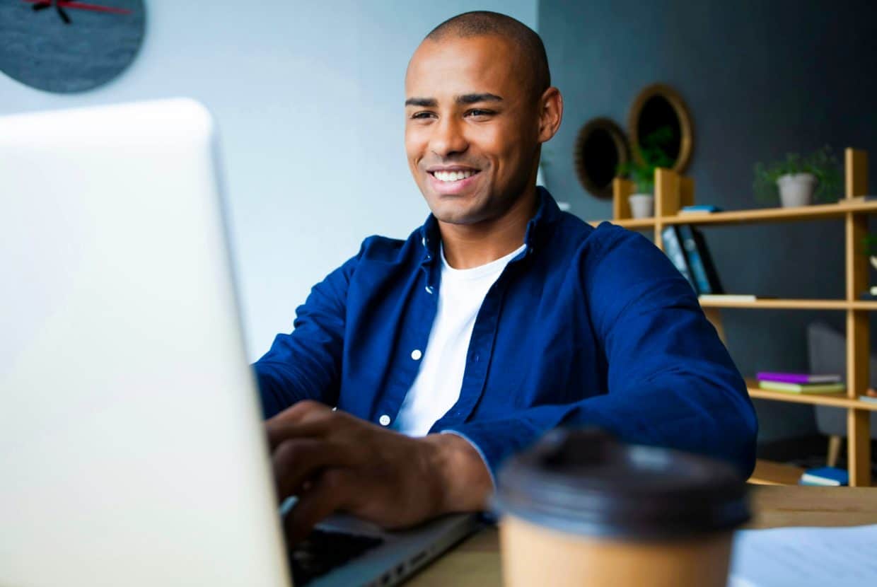 Man smiling while working on a laptop at a desk with a coffee cup nearby.
