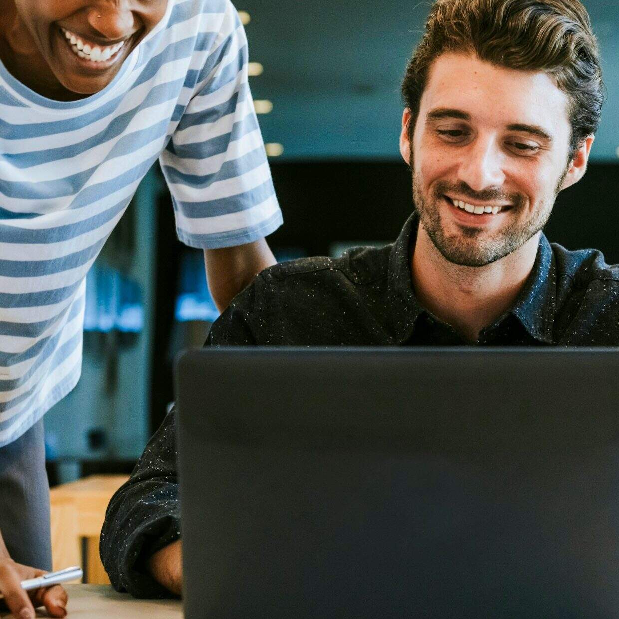 Two co-workers smiling while looking at a laptop screen in a modern office.