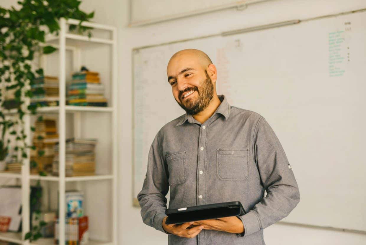 Smiling man holding a tablet, with a whiteboard and bookshelves in the background.