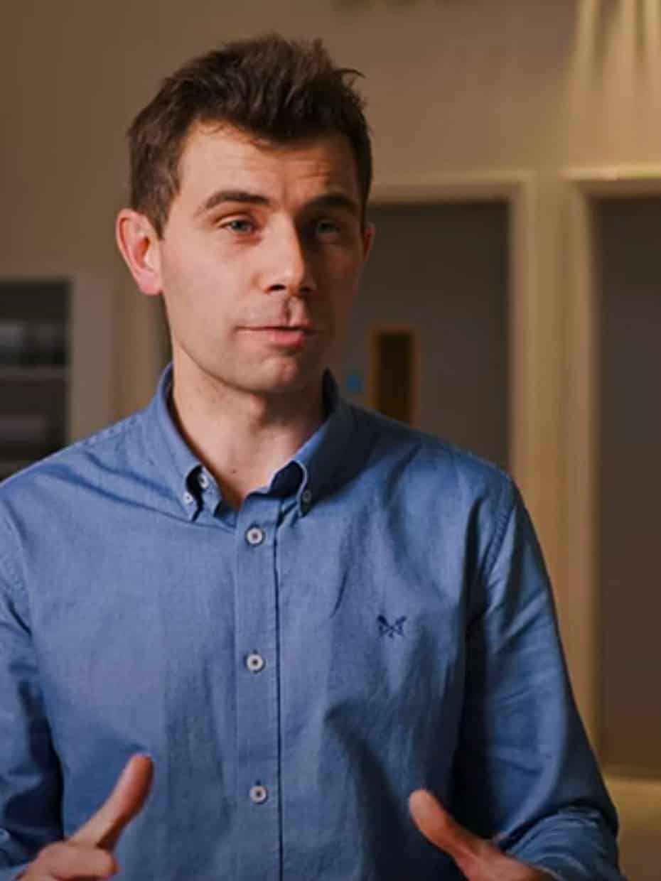 Man wearing a blue shirt speaking indoors, gesturing with his hands in front of closed double doors and soft lighting.
