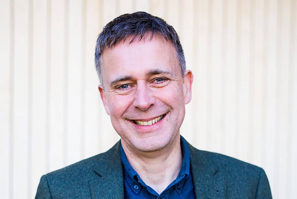 Smiling man wearing a dark green blazer and blue shirt, standing in front of a light vertical panel background.