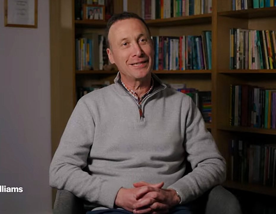 Man sitting and smiling in a chair in front of bookshelves filled with books, with text overlay in the corner reading 'Dyfan Williams, Pastor'.