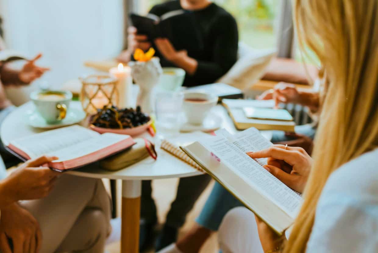 A group of people sitting around a table reading Bibles, with coffee, fruit, and candles on the table, suggesting a relaxed Bible study setting.