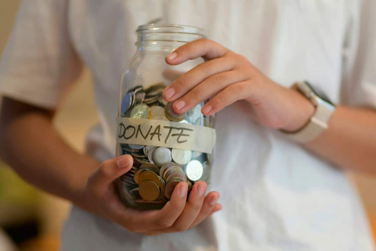 Person holding a glass jar filled with coins labeled 'Donate'.