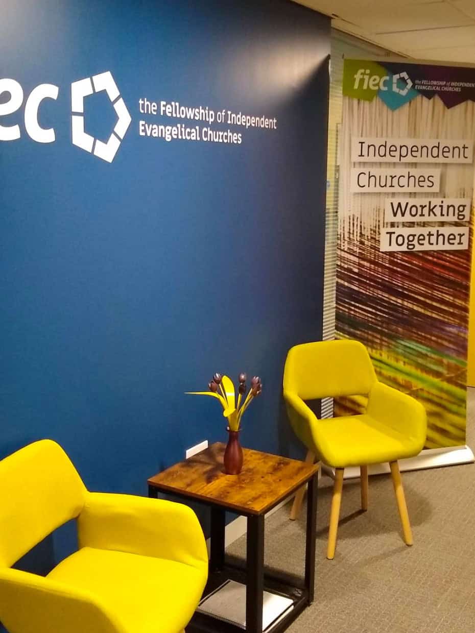 Modern office reception area with two yellow chairs, a small table, and a blue wall displaying the Fellowship of Independent Evangelical Churches logo.
