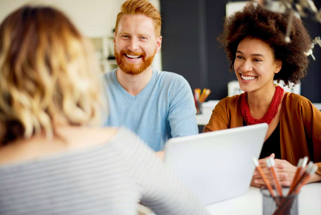 Smiling couple sitting at a desk with a laptop, talking to a person facing them in an office setting.
