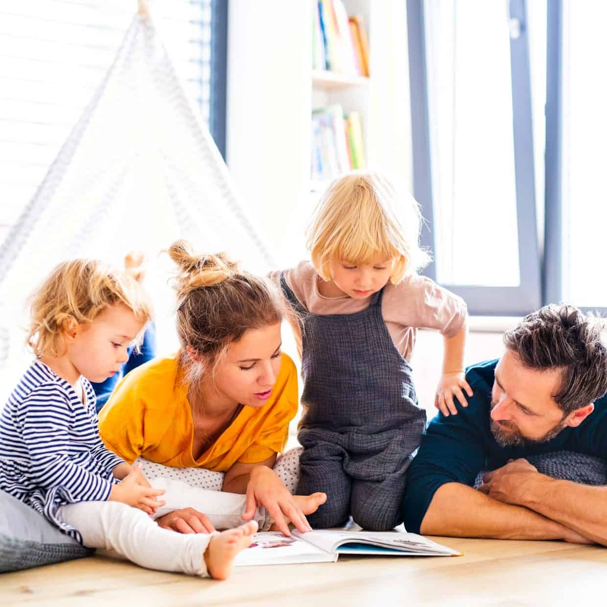 Young family lying on the floor reading a book together in a bright living room.