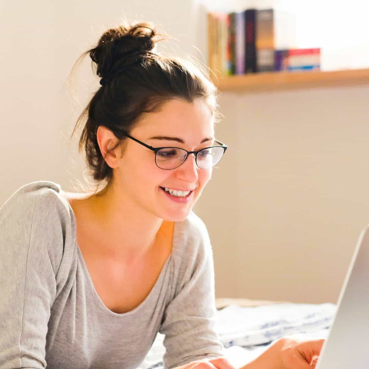 Smiling woman lying on a bed using a laptop in a sunlit room.