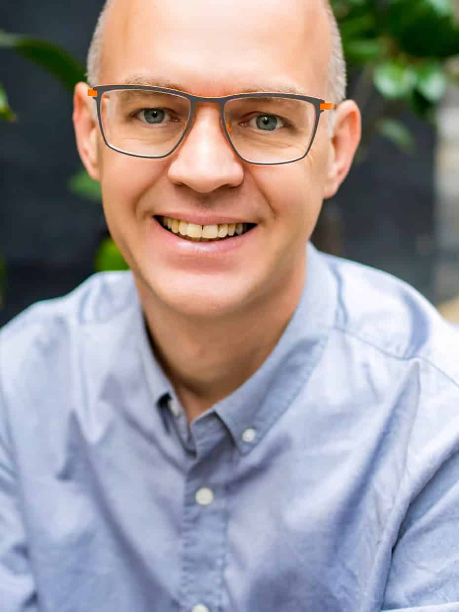 Smiling man wearing glasses and a light blue shirt, seated indoors with green plants in the background.