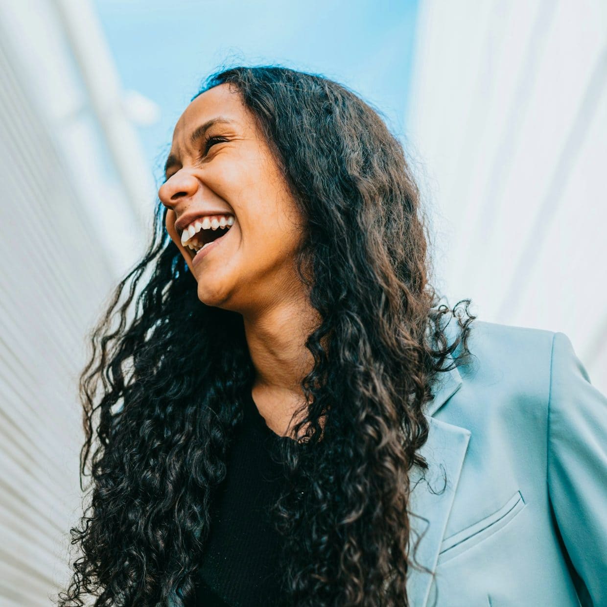 Joyful woman with long curly hair laughing outdoors in sunlight.