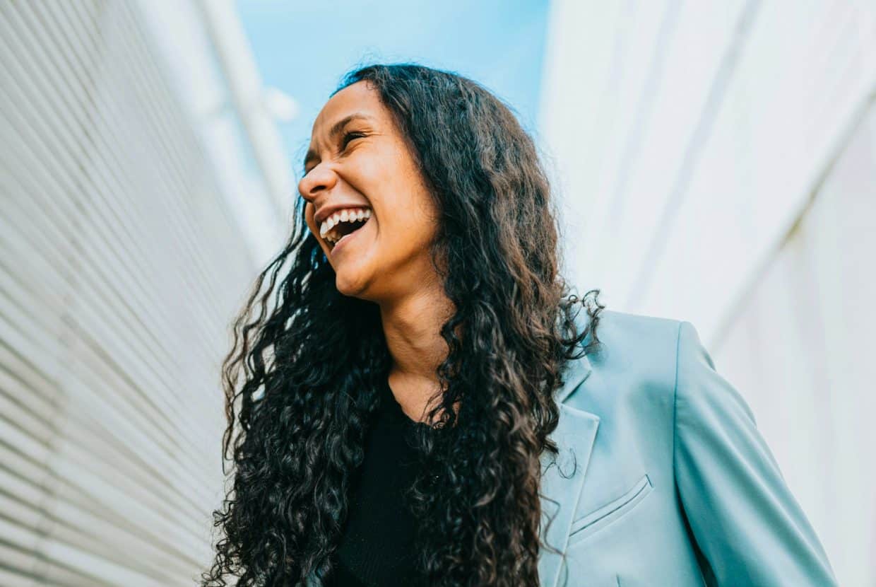 Joyful woman with long curly hair laughing outdoors in sunlight.
