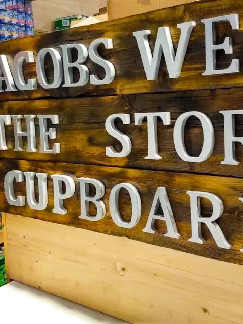 Wooden sign reading “Jacobs Well The Store Cupboard” displayed inside a food storage area with shelves of packaged goods in the background.