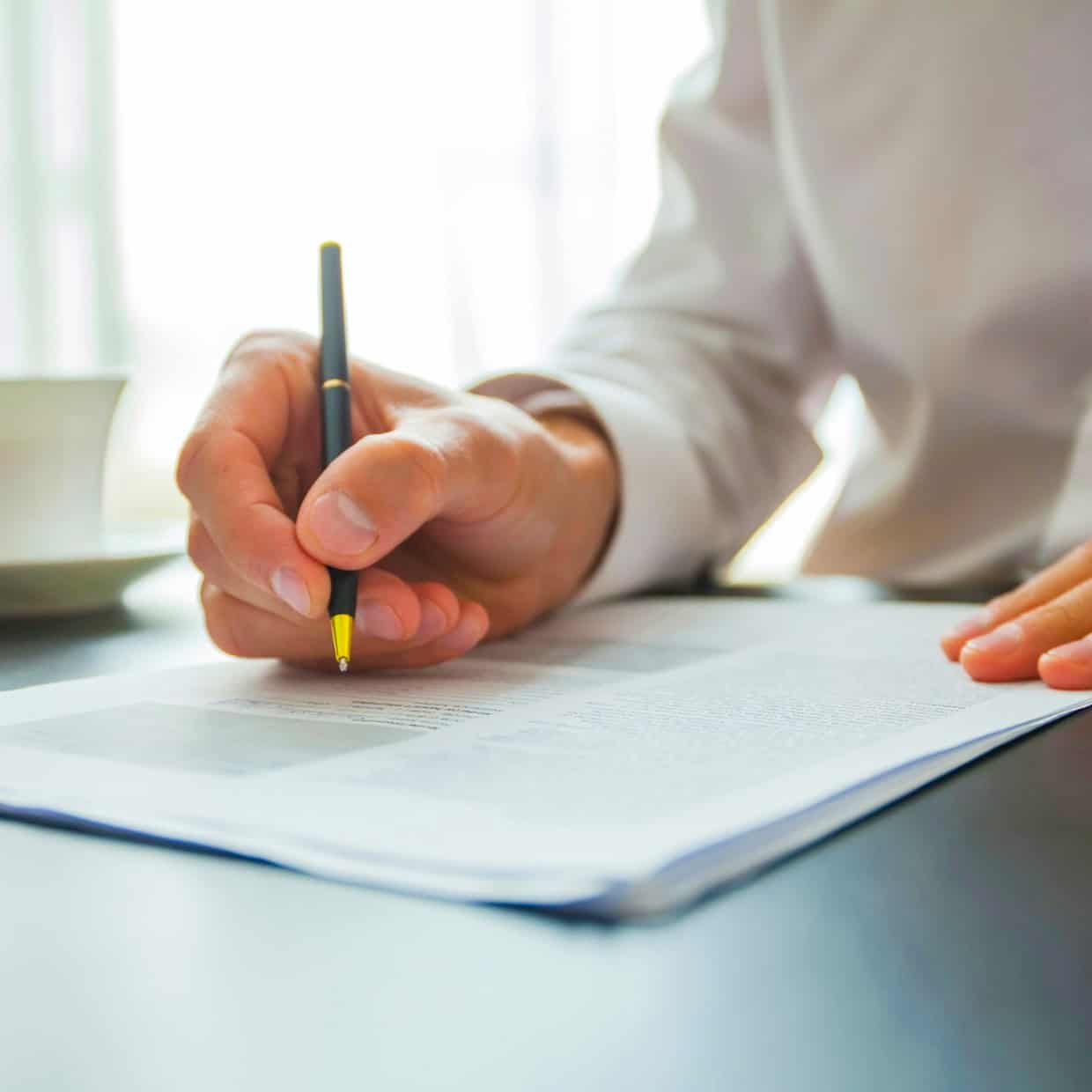 Person in a white shirt signing a document at a desk with a cup and saucer nearby.