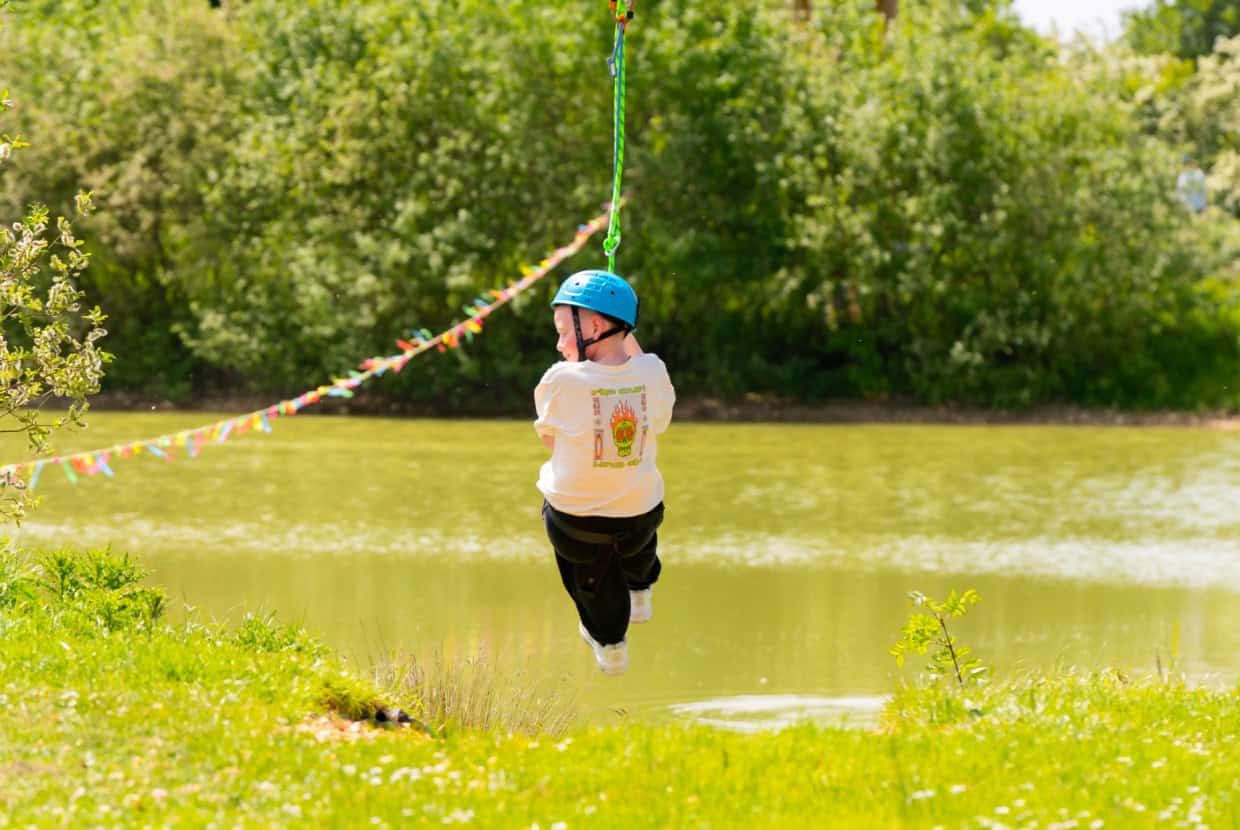 Child wearing a blue helmet and white T-shirt riding a zip line over a small lake surrounded by green trees and grass.