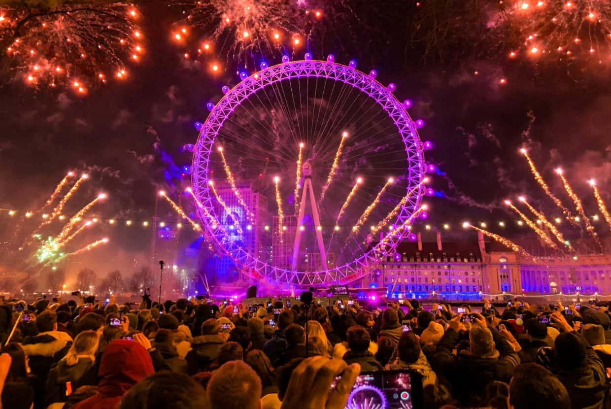 Large crowd watching fireworks bursting around the illuminated London Eye at night during New Year celebration.