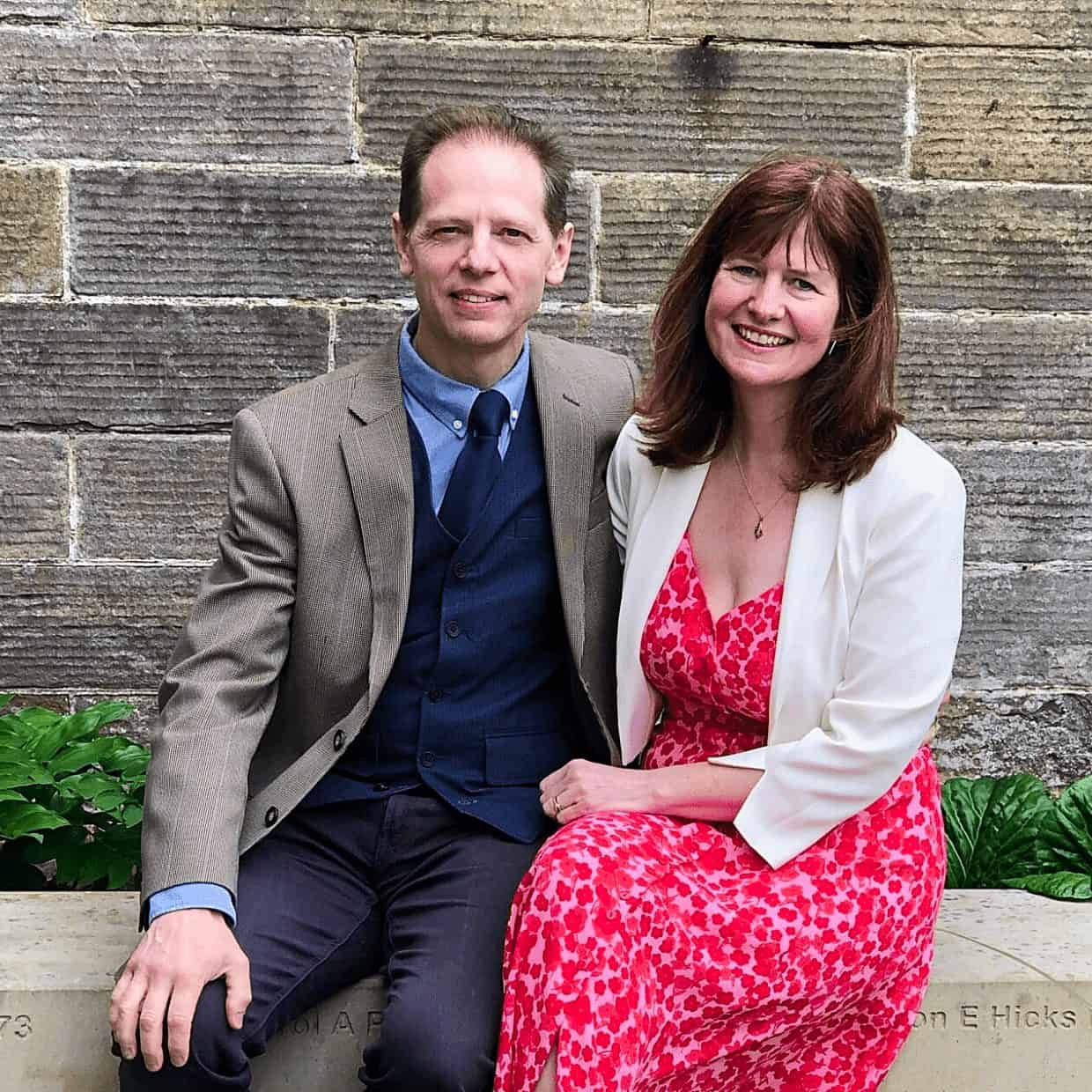 Smiling couple sitting together on a stone bench in front of a brick wall, dressed smartly in a suit and a pink floral dress.