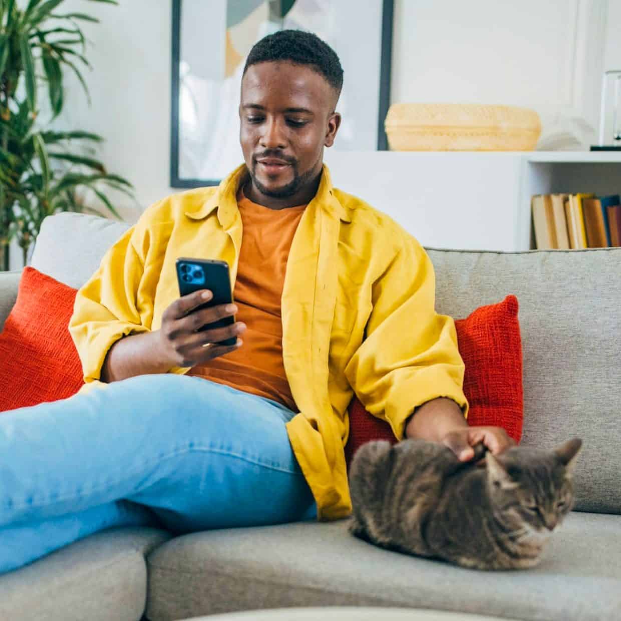Man relaxing on a sofa, using his phone and petting a cat in a cosy living room.